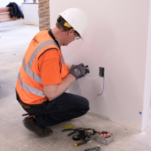 Electrician wearing a white helment and other protective gear fixes outlet wiring.