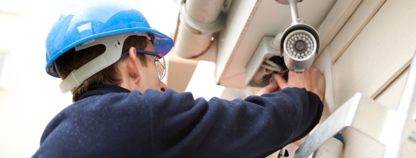 An electrician in a blue sweatshirt and blue helmet installs a building's security lighting.