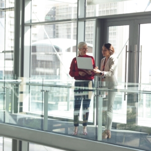 Two people stand in an office building that has large windows.