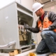 Male electrician in white hard hat and orange safety vest works on a commercial business's electrical system.