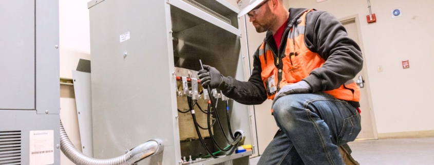 Male electrician in white hard hat and orange safety vest works on a commercial business's electrical system.
