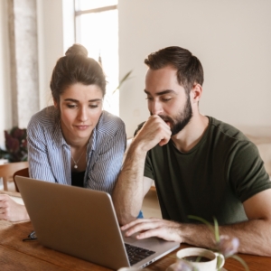 Man and woman sit at kitchen table and scroll through websites on a laptop.