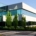 White, modern office building with wraparound windows near a stone-paved walkway, and green trees and bushes, with blue sky above.