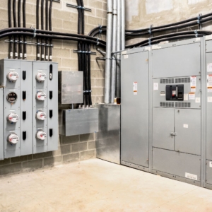 Large, grey metal electrical machinery and many black cables in a warehouse with white stone floor and walls.