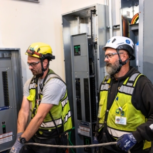 Two commercial electricians with beards in grey and black shirts fix data cables in a warehouse.