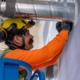 Male commercial electrician with safety goggles, yellow hardhat, blue gloves, and orange shirt stands on blue ladder while fixing a warehouses conduit system.