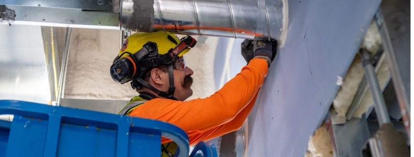 Male commercial electrician with safety goggles, yellow hardhat, blue gloves, and orange shirt stands on blue ladder while fixing a warehouses conduit system.