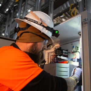 Commercial electrician with white helmet, orange shirt, and blue gloves fixes a complex electrical panel in a warehouse.