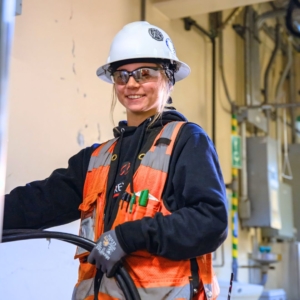 Female electrician in white hardhat wears safety goggles and orange safety vest with a blue sweatshirt underneath it and holds cables.