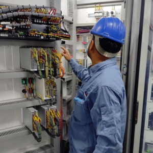 Commercial electrician wears a blue safety helmet and blue work shirt while working on a complex electrical panel with many multicolored cables.