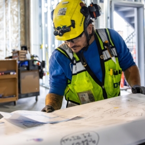 Industrial electrician wearing a yellow hard hat and blue t-shirt, reviewing engineering plans inside a building.
