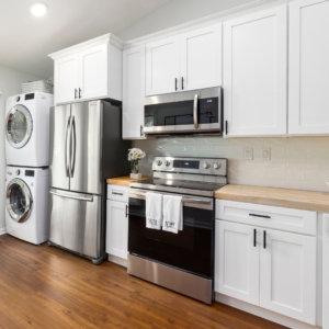 Modern kitchen with white cabinets, stainless steel fridge and stove, stackable washer and dryer, hardwood floors, and wood countertops.