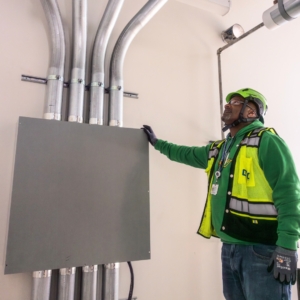 Industrial electrician wearing a green hard hat and sweatshirt, examining wiring panels and conduit.
