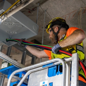 Industrial electrical contractor wearing an orange shirt and yellow safety vest, installing wiring in a ceiling.