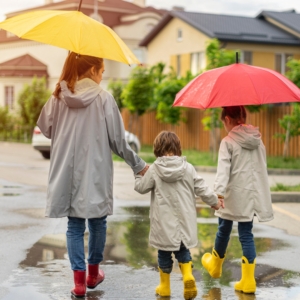 Three kids in grey raincoats walk down the street with yellow and red umbrellas.