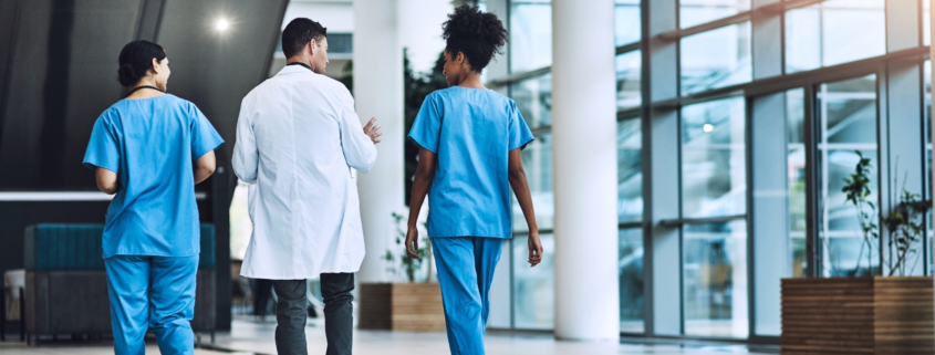 Three doctors walking down a hospital corridor with large windows.