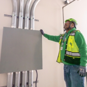 Industrial electrician in green sweatshirt near electrical panel with conduit.