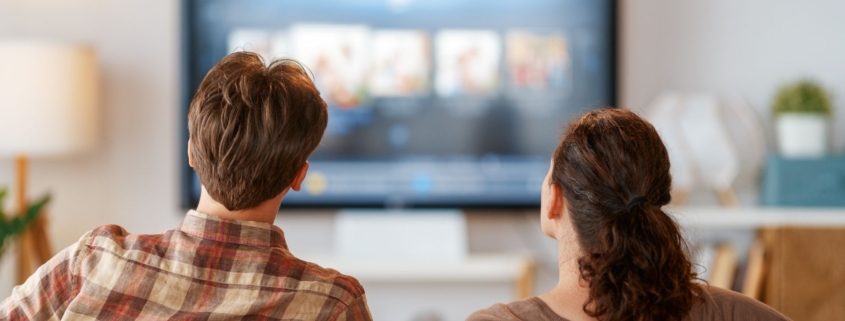 Man and woman sitting on a gray couch watching TV in their living room.