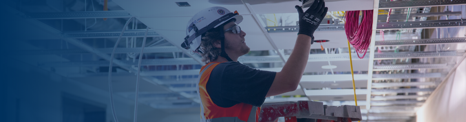 An IBEW electrician in a white hard hat and safety vest standing on a ladder routing red and yellow cables through an overhead cable tray in a commercial building.