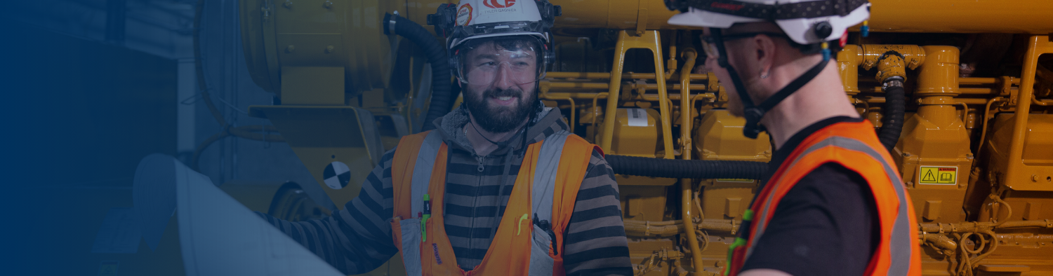 Two IBEW electricians in white hard hats and orange safety vests smiling and talking in front of a large yellow industrial generator.