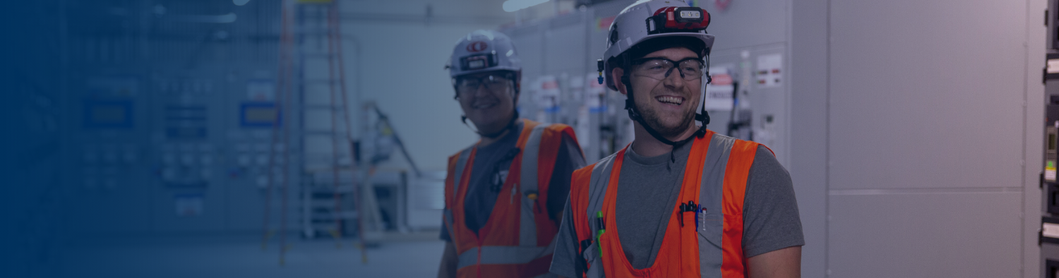 ibew-electricians-smiling-electrical-facility Two smiling IBEW electricians in white hard hats, safety glasses, and orange high-visibility vests inside an electrical facility.