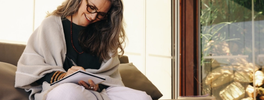 Smiling woman with glasses and a shawl writing in a notebook while sitting on a couch in a sun-filled living room.
