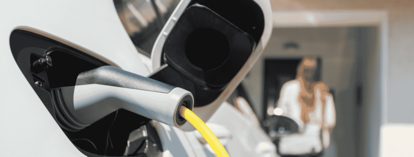 Close-up of an electric vehicle charging port with a yellow cable plugged in, with a person blurred in the background.