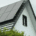 Solar panels installed on the sloped roof of a white residential home surrounded by green trees on a cloudy day.