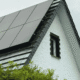 Solar panels installed on the sloped roof of a white residential home surrounded by green trees on a cloudy day.