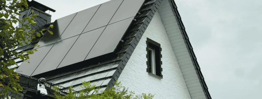 Solar panels installed on the sloped roof of a white residential home surrounded by green trees on a cloudy day.