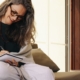 Smiling woman with glasses and a shawl writing in a notebook while sitting on a couch in a sun-filled living room.