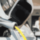 Close-up of an electric vehicle charging port with a yellow cable plugged in, with a person blurred in the background.
