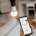A person controlling a smart home lighting app on a smartphone next to an illuminated light bulb in a modern home.