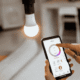 A person controlling a smart home lighting app on a smartphone next to an illuminated light bulb in a modern home.