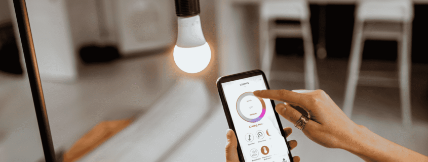 A person controlling a smart home lighting app on a smartphone next to an illuminated light bulb in a modern home.